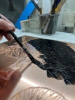 Maler en kobberplade med sort maling og pensel.